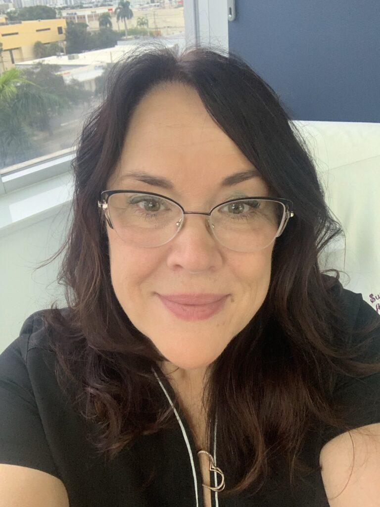A woman with wavy dark hair and glasses smiles at the camera. She is wearing a black top with white piping and is seated indoors near a window with a cityscape and greenery in the background.