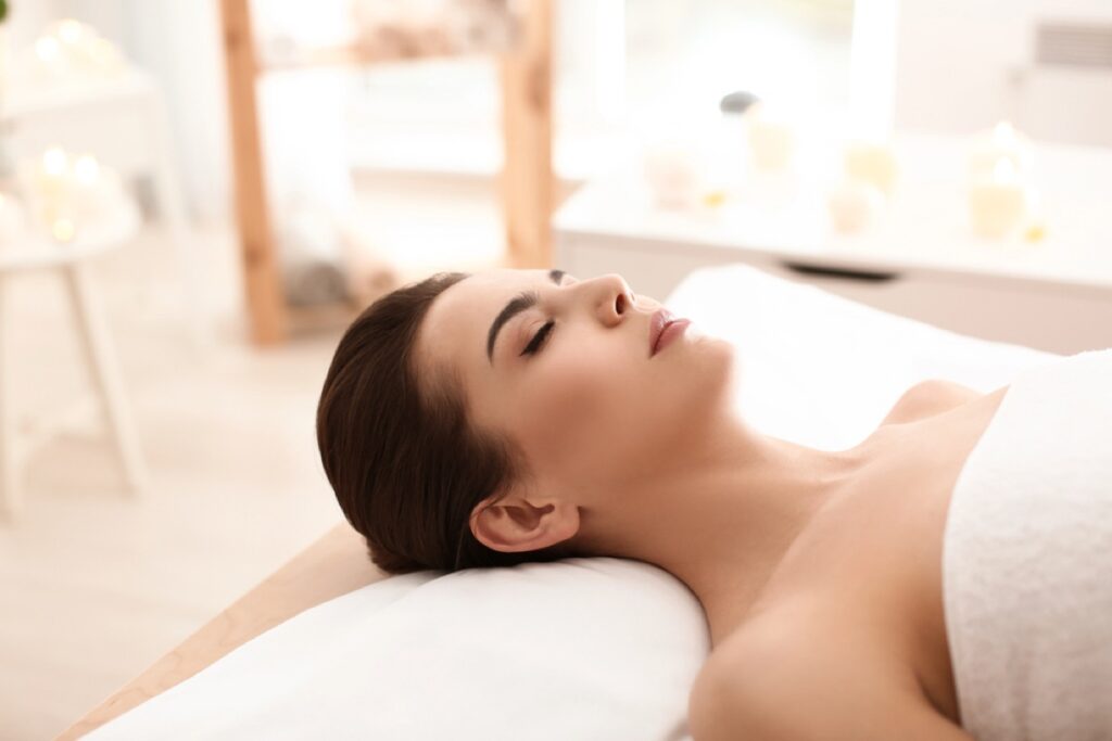 A woman with dark hair wrapped in a towel is lying on her back with eyes closed, relaxing in a spa setting with soft lighting and blurred candles in the background.
