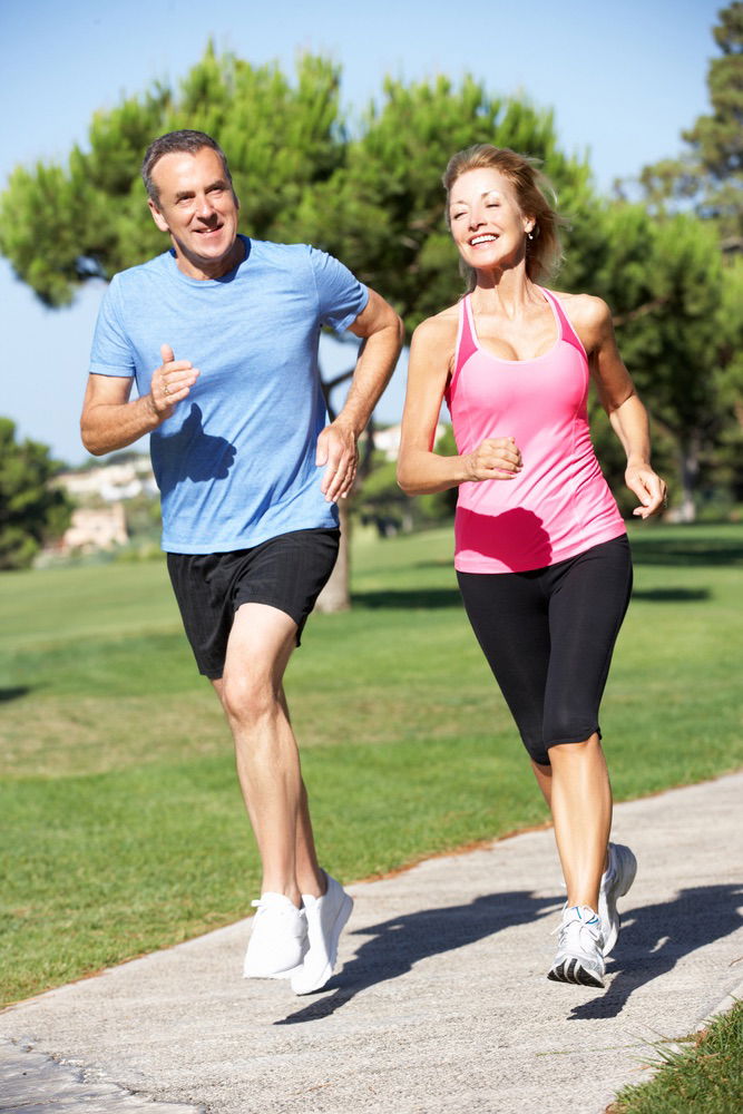 A man and a woman jog together on a park path, smiling and wearing athletic clothes. Trees and grass are in the background on a sunny day.