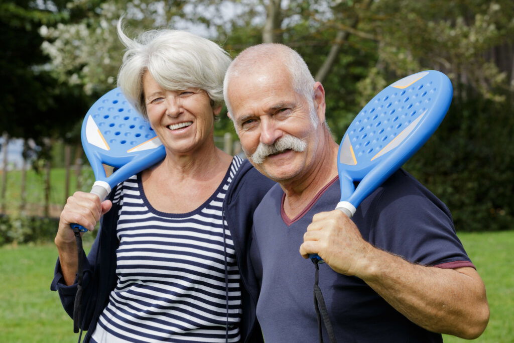Smiling older couple standing outdoors, each holding a blue paddle over their shoulder, suggesting they are about to play a paddle sport. They appear happy and relaxed in a green, sunny park—enjoying life with the help of integrative medicine.