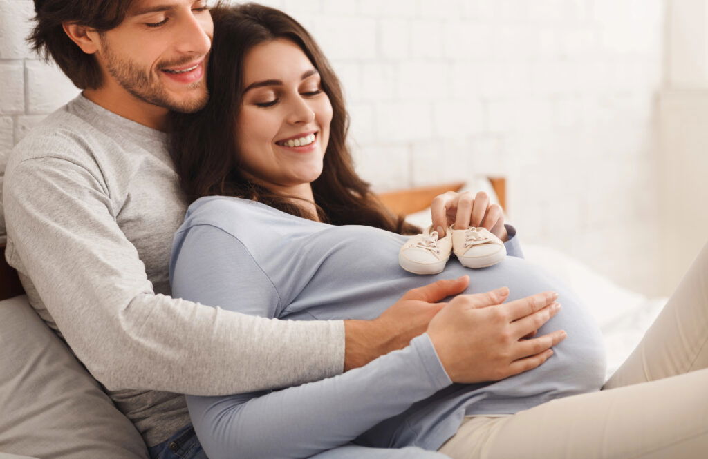 A smiling pregnant woman sits with a man who embraces her from behind. She holds tiny baby shoes on her belly while they both look happy and content, sitting together in a bright, cozy room.