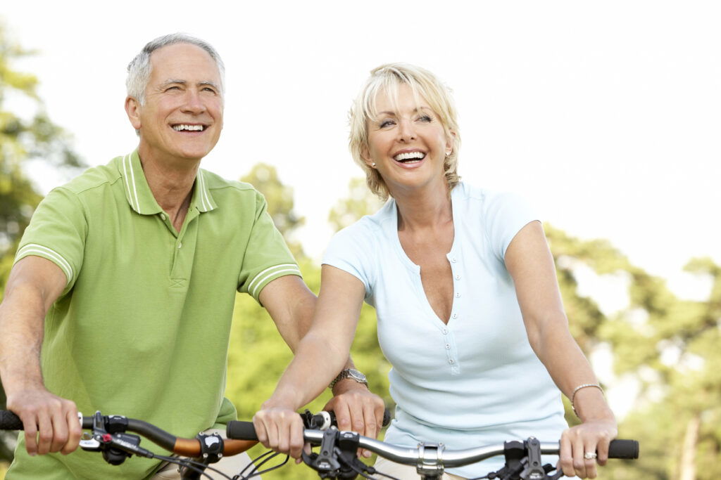 Two smiling older adults, a man and a woman, ride bicycles outdoors on a sunny day. They appear happy and relaxed, wearing casual clothes, with trees and a bright sky in the background.